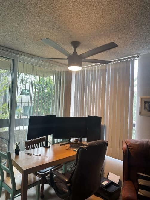 Home office setup featuring dual monitors, a wooden desk, and a ceiling fan with natural light.