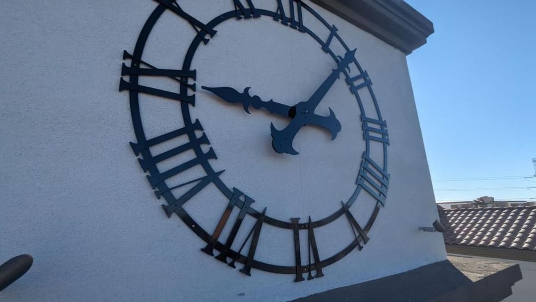 Black metal clock on a white wall with Roman numerals against a clear blue sky.