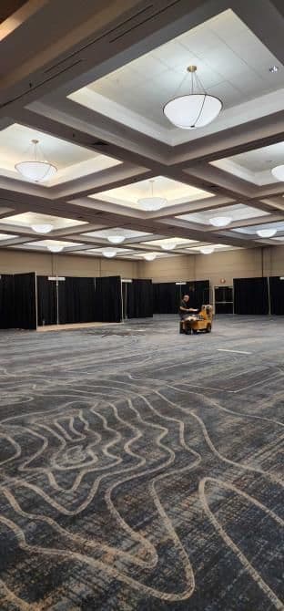 Empty event space with carpet markings and a worker operating a floor cleaning machine.